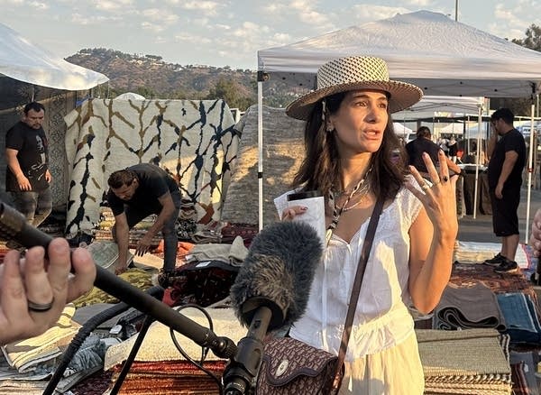 A person holds a mic toward a man and a woman talking. The woman, center, is wearing a white top and cream-colored pants and sunhat. The man is older, with gray hair and a maroon shirt. They're standing in a parking lot turned into a flea market, where vendors are displaying rugs and art behind…