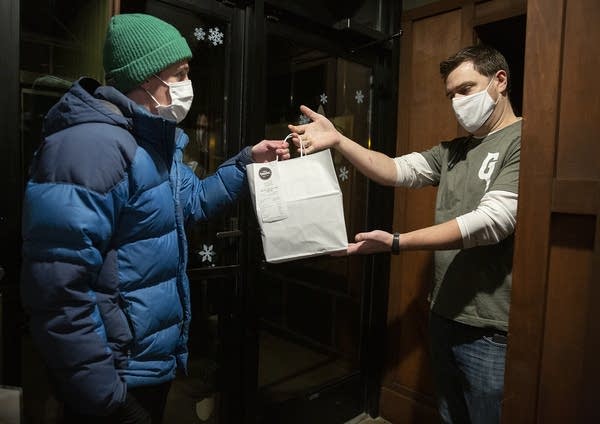 A man wearing a mask hands another person a bag of food. 