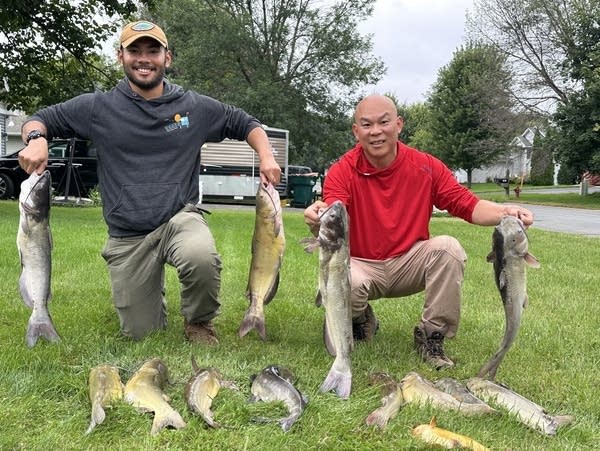 Two men smile and hold up four large fish, with more fish laid on the ground