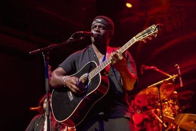 A musician sings and plays guitar onstage in a large music venue