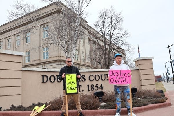 Two people hold signs outside of a county courthouse.