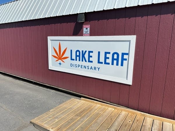 A sign for "Lake Leaf Dispensary" on a maroon wall. It has blue text on a white background next to an orange cannabis leaf.