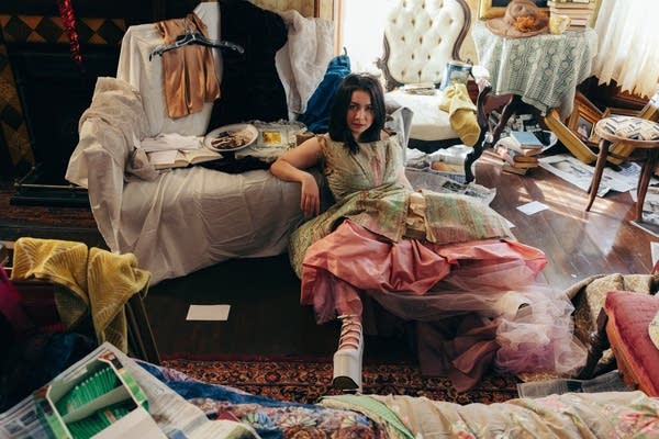 A woman sits on the floor of a room cluttered with bric-a-brac evoking the Victorian era
