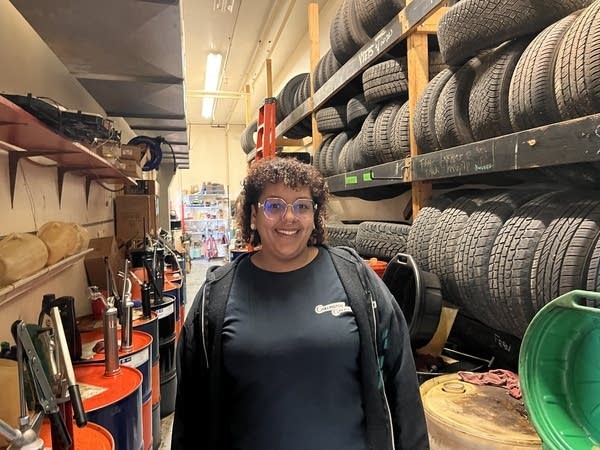 A woman stands in front of tires and oil drums in an auto shop.