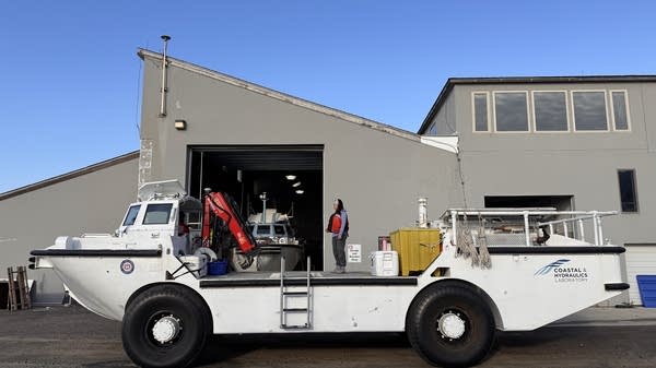 A white amphibious vessel, part boat, part truck, sits on concrete in front of a low building.