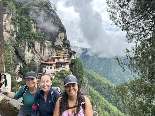 Three smiling women wearing backpacks stand next to each other. Behind them is structure nestled up against a steep wall of rock with trees growing on it. Below them is a plunging valley covered in greenery.