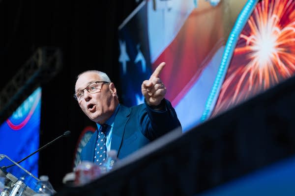 A man speaks on a stage at the convention, pointing out across the crowd.