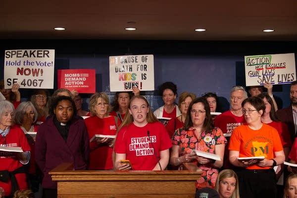 A large group stands with red shirts and signs while one woman speaks into a microphone.