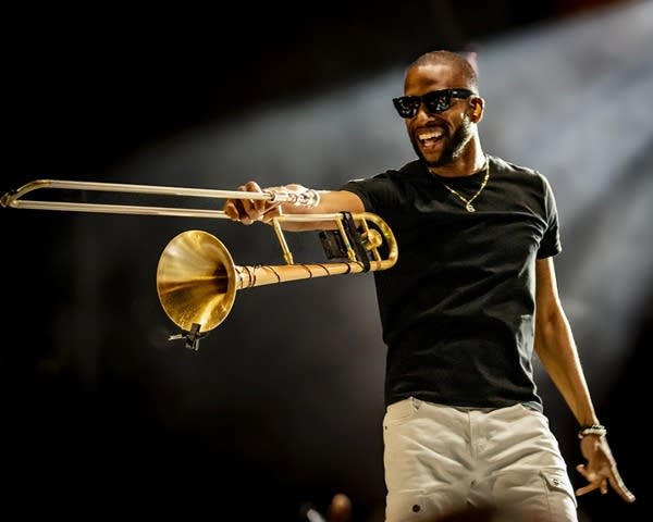 A man in sunglasses smiles and holds out a trombone