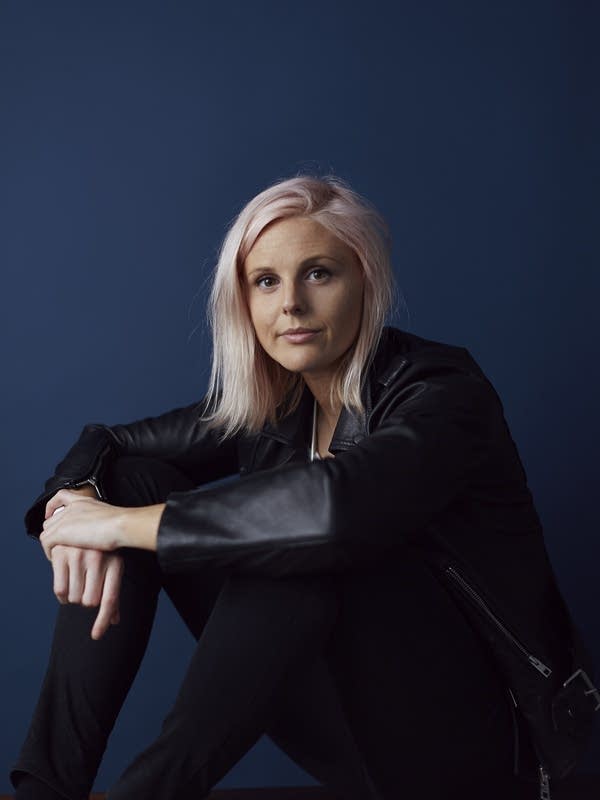 A young blonde woman in a black suit sits in front of a navy blue wall.
