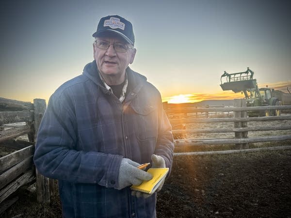 A man in a ball cap holds a yellow notepad. The sun rises behind him.