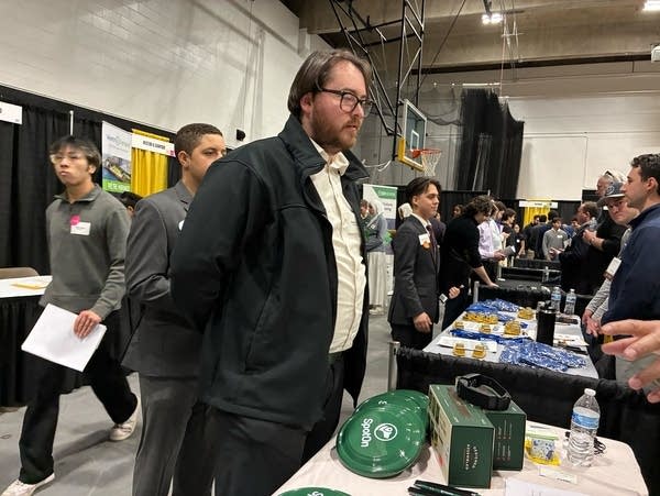A man in a white button-up and black jacket stands at a career fair table.