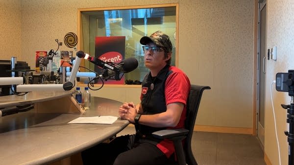 A man sits at a table in a broadcast studio and speaks into a microphone
