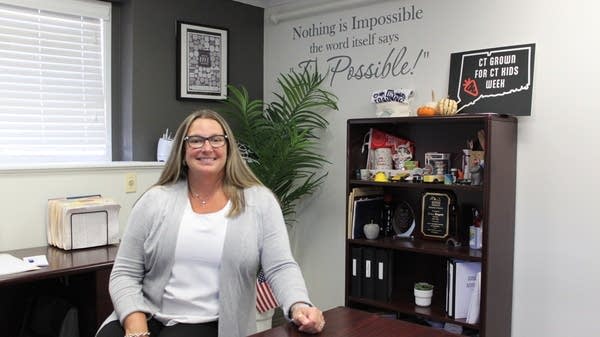 Erica Biagetti sits at her desk and smiles to the camera. Behind her, a shelf contains signs, including ones that say "CT Grown for CT Kids Week." On the wall, a decal reads: "Nothing is impossible. The word itself says I'm Possible!"