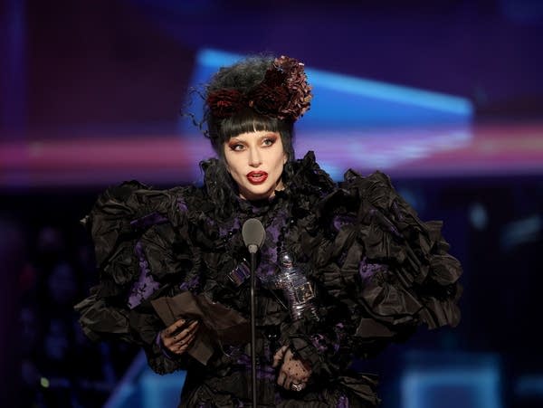 Lady Gaga wears a black dress and speaks into a microphone.