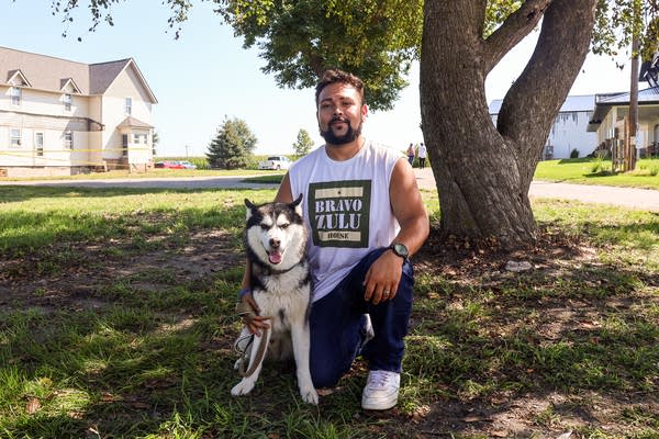 A man poses for a photo and kneels down with his husky under a tree.