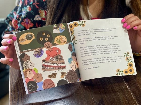 A woman holds open a children's book. The left page has an illustration of a girl speaking to other children.
