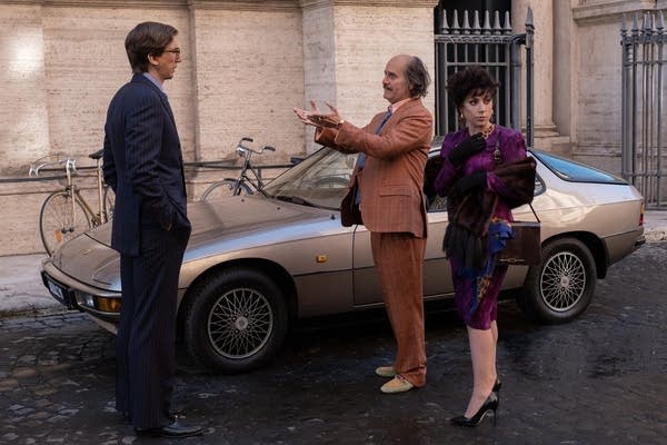 Three people interact next to a parked car on a street in Italy