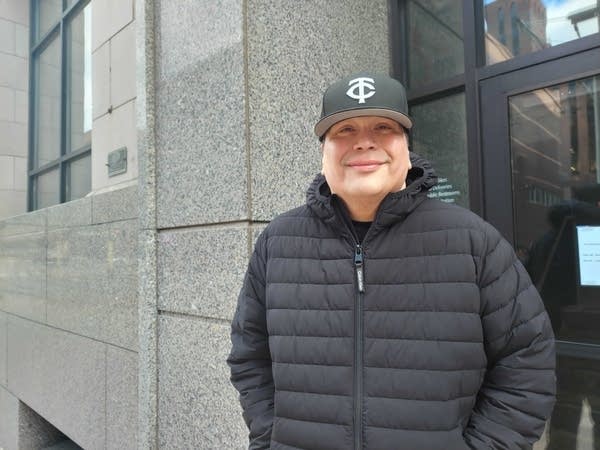 A man in a black baseball cap and black coat poses for a photo in front of a gray wall.