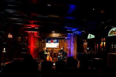 Two musicians sing while playing cello and guitar in a pub
