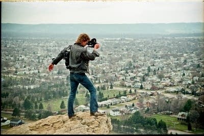 A man holds a film camera and looks over a town from atop a bluff.