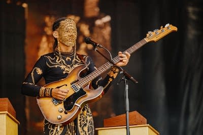 A musician in a golden mask plays bass on a large outdoor stage