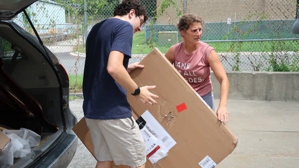 Homes Not Borders executive director Laura Thompson Osuri, , wearing a pink t-shirt with rolled up sleeves, moves a large cardboard box containing a bedframe with the help of her nephew, Jack Campbell, who is wearing a blue t-shirt and khaki shorts. He is tall and has to duck under the open door of…