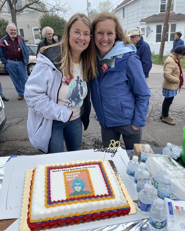 Two women pose for photo behind a cake decorated with Bob Dylan's face.