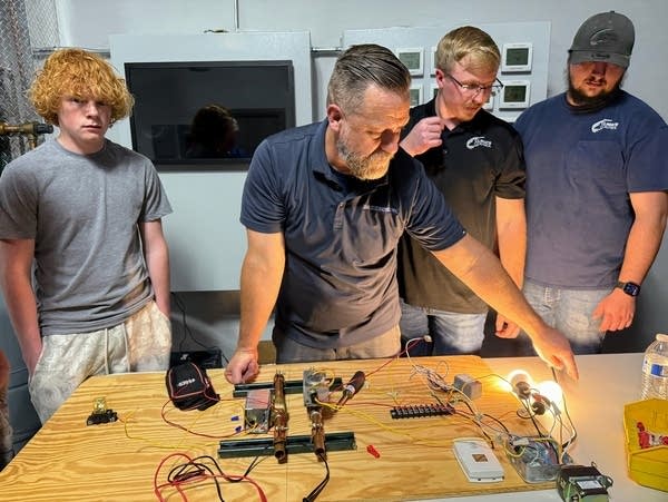 A man with a beard points to a light bulb while students look on.