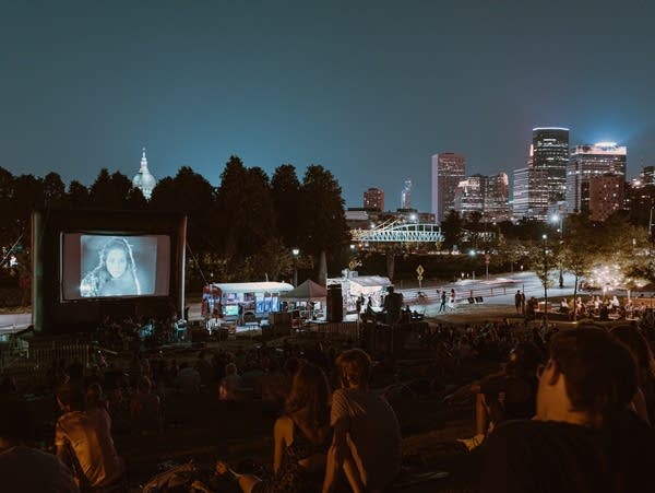 People watch a film on an outdoor screen with the Minneapolis Skyline in the background.