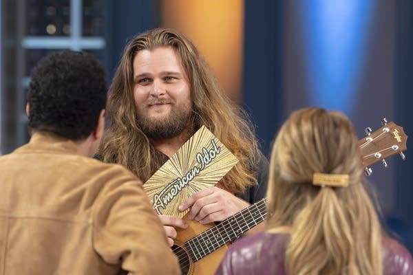 A singer with a guitar holds an "American Idol" golden ticket in front of the judges.