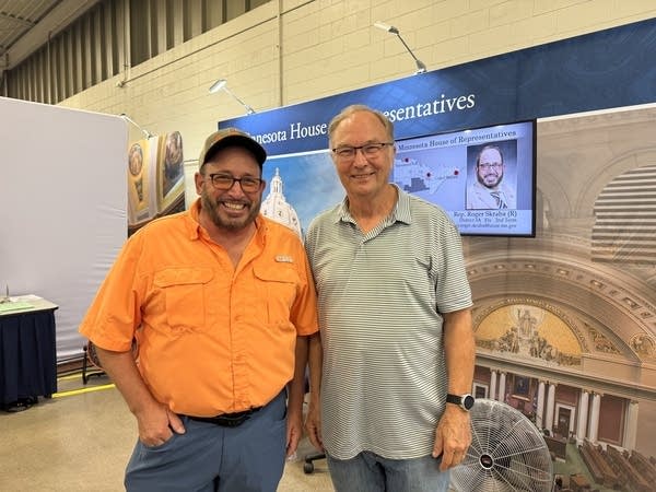 A man in a bright orange shirt and baseball cap, left, poses with a man in a green polo shirt.