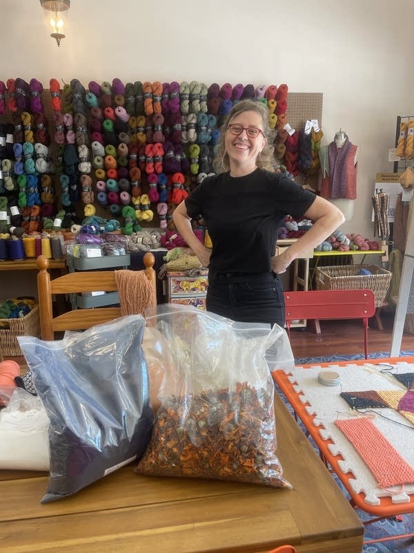 Bags of indigo and marigold sit on a wooden table. A woman in a black shirt stands behind them, with a colorful array of yarn in the background.