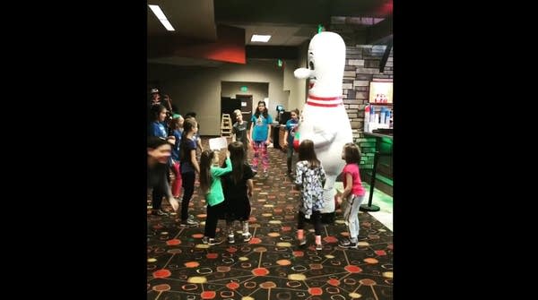 A bowling pin mascot surrounded by a entertained kids at bowling alley