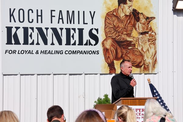 A man speaks into a microphone at a podium in front of a sign reading, "Koch Family Kennels: For loyal & healing companions."