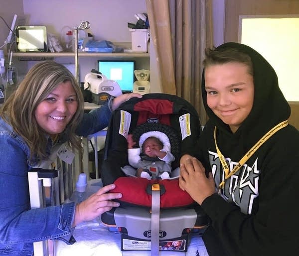 A young teen and woman smile with a baby in a carseat.