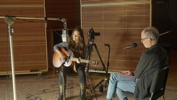 A woman sings and plays guitar in a recording studio