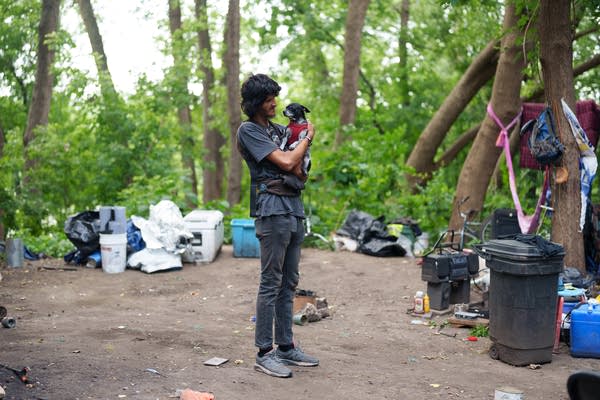 A man holds his dog in the middle of an encampment in the woods.