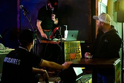 Justin Vernon wears a black shirt and cap at a bar and speaks into a microphone.