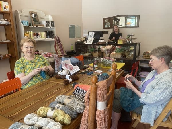 Two women sit knitting at a wooden table in a yarn store in Baltimore. 