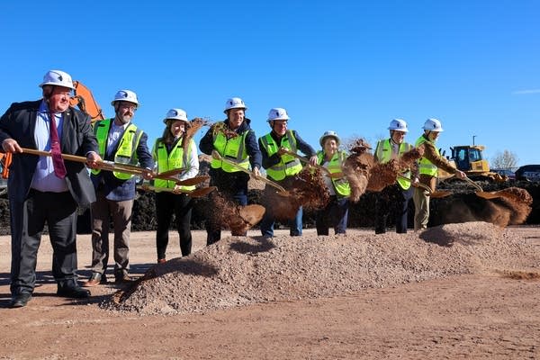 A group of people in green hi-vis vests and hard hats dig up dirt with shovels.