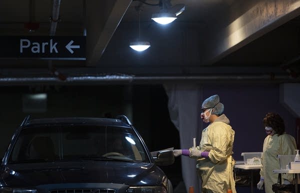 A person wearing a face mask and PPE talks to a person in a car. 