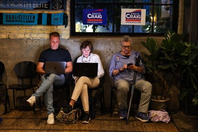 People in chairs against a wall look at their laptops and phone.