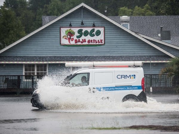 A small van drives through floodwater in front of a restaurant