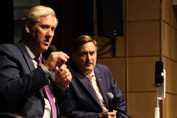 A man in a suit on the left speaks into a microphone while another man in a suit leans in to listen.