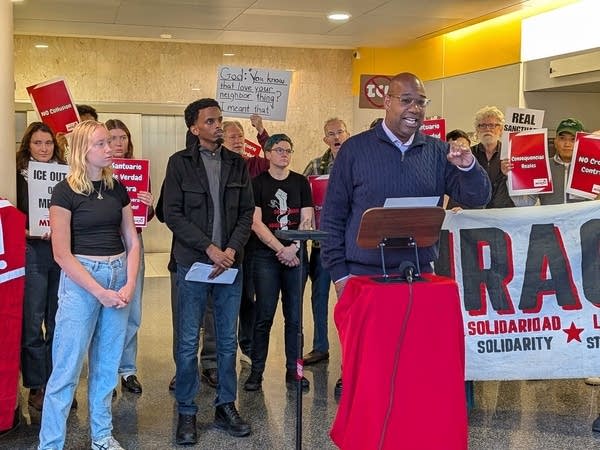 A man speaks in a building lobby surrounded by people holding red and white signs.