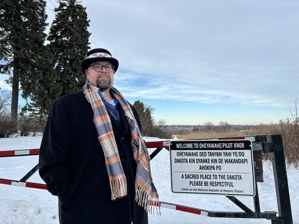 A man stands next to a gate while wearing a winter coat, hat and scarf.