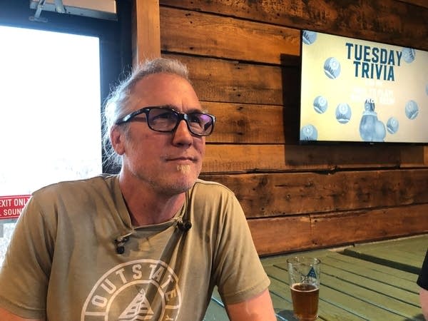 A man in a tan shirt and glasses looks on while a TV mounted on the wall behind him says "Tuesday Trivia."