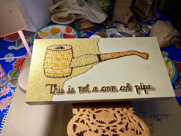 An in-progress work of crop art lays on a craft table. It shows a pipe made of seeds with the text, "This is not a corn cob pipe."