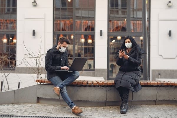 Two people with masks on sit on a bench outside